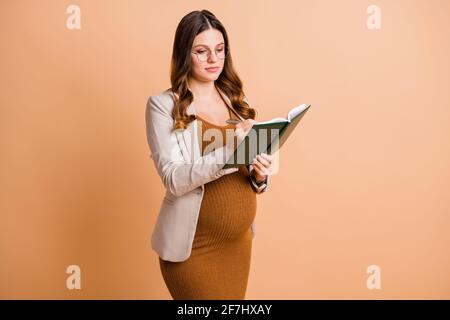 Portrait de la jolie femme enceinte intelligente et intelligente qui apprend à écrire quotidiennement note hebdomadaire isolée sur fond beige pastel Banque D'Images