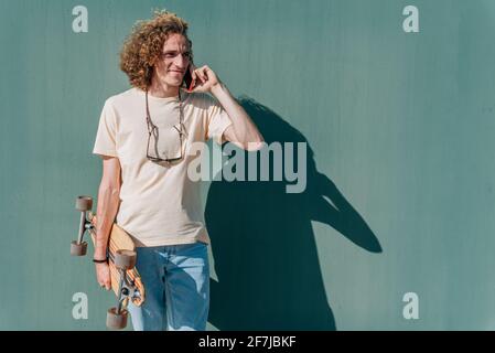 portrait horizontal d'un jeune homme aux cheveux longs parlant au téléphone. Il porte des vêtements décontractés et tient une planche à roulettes Banque D'Images