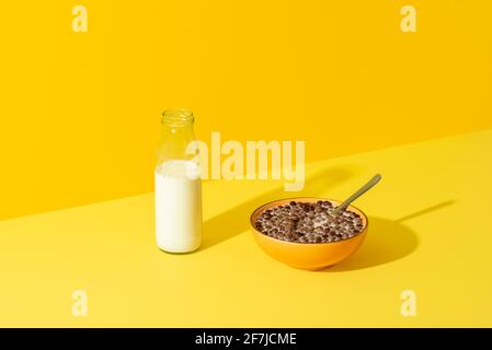 Bol avec céréales et une bouteille de lait en verre sur une table jaune, en lumière vive. Table de petit déjeuner avec un bol à céréales et une bouteille de lait. Banque D'Images