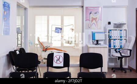 Salle d'attente dans la clinique avec personne dans elle, le bureau principal avec nouvelle normale ayant signe sur la chaise pour la distance sociale en cas de pandémie de coronavirus. Réception de stomatologie vide pendant l'épidémie de covid-19. Banque D'Images