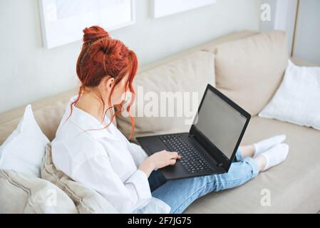 Une fille aux cheveux rouges et brillants est assise sur le canapé avec ordinateur portable. Banque D'Images