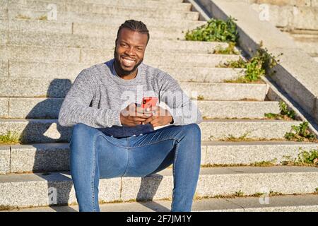 Homme afro-américain assis dans les escaliers souriant, regardant la caméra, un jour ensoleillé. Joyeux jeune Latino homme. Photo de haute qualité Banque D'Images