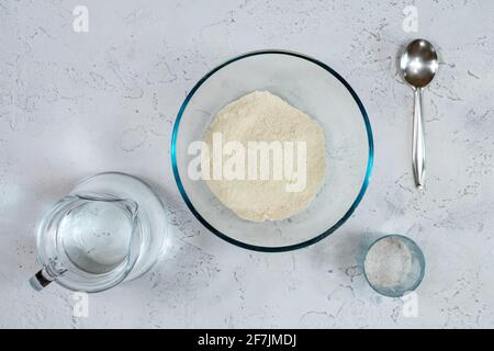 Bols en verre et en céramique avec farine de blé, verre et verseuse d'eau pour pétrir la pâte sur une table blanche. Banque D'Images