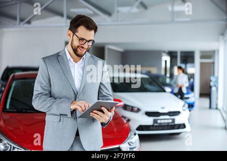 Vendeur de voiture souriant et amical se tenant dans un salon de voiture et utilisant une tablette pour vérifier les nouveaux messages que les clients publient sur Internet. Banque D'Images