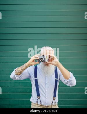 Joyeux homme hipster à l'aide d'un vieux appareil photo vintage debout sur le mur rouge - un photographe mûr et élégant qui s'amuse à voyager dans le monde - joyeuse vie de personnes âgées Banque D'Images