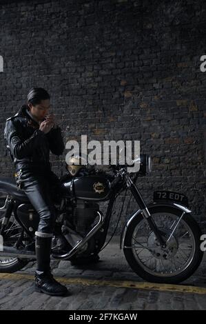 biker , coureur de café portant une veste en cuir noir assis sur une moto britannique classique et fuyant une cigarette à Londres, Royaume-Uni Banque D'Images