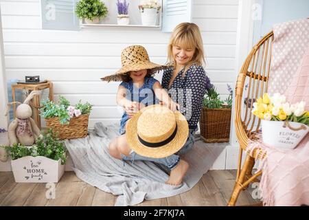 petite fille de 4-5 ans joue avec des chapeaux de paille sur la véranda de la maison avec la mère dans ses bras. Rire, bonheur, joie, concept de maternité Banque D'Images