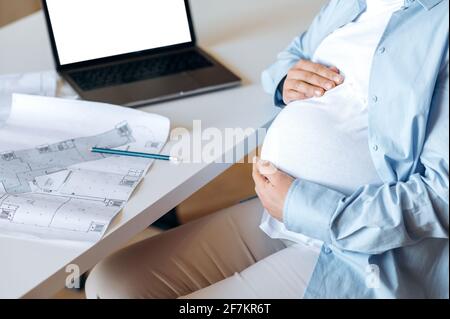 Une femme enceinte, designer ou pigiste, s'assoit à un bureau, épouse doucement son ventre avec ses mains, sur la table un ordinateur portable avec un écran blanc vierge et un document de travail Banque D'Images