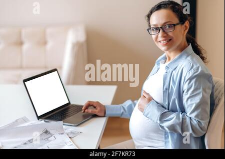 Une femme enceinte, designer ou pigiste, s'assoit à un bureau, épouse doucement son ventre avec sa main, regarde et sourit dans l'appareil photo, sur la table un ordinateur portable avec écran blanc vierge et des documents de travail Banque D'Images