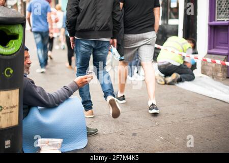 Un pauvre âgé mendiant dans une rue haute, tandis que deux garçons négligents passent devant et un travailleur occupé en arrière-plan. Camden High St, Londres, Royaume-Uni. Août 2015 Banque D'Images