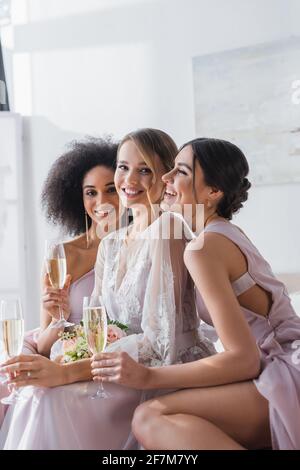 bonne mariée regardant l'appareil photo près des demoiselles multiculturelles avec du champagne verres dans la chambre Banque D'Images