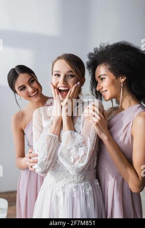 émerveillement de la mariée en touchant le visage tout en regardant l'appareil photo près de multiculturel les bridesmaides Banque D'Images