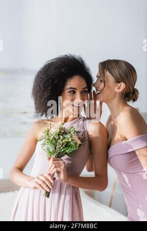 une femme joyeuse qui dit secret à un ami afro-américain souriant tenant bouquet de mariage Banque D'Images