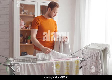 Homme faisant des tâches à la maison. L'homme caucasien enlève les vêtements et les draps de bébé après la lessive du sèche-linge portable dans le salon Banque D'Images