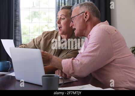 Surpris couple âgé caucasien dans la salle de séjour à la table en utilisant un ordinateur portable, payer des factures et parler Banque D'Images