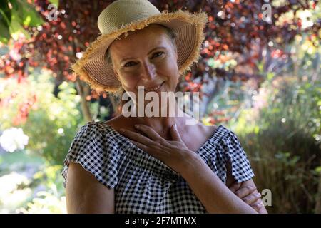 Portrait d'une femme âgée caucasienne heureuse debout dans un jardin ensoleillé portant un chapeau de soleil, souriant à l'appareil photo Banque D'Images
