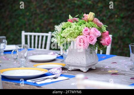 Table dans le jardin avec des assiettes en porcelaine bleue et jaune, avec des verres en cristal vides et un arrangement de fleurs au centre avec des roses roses roses et blanches Banque D'Images