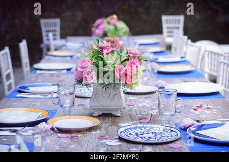 Table dans le jardin avec des assiettes en porcelaine bleue et jaune, avec des verres en cristal vides et un arrangement de fleurs au centre avec des roses roses roses et blanches Banque D'Images