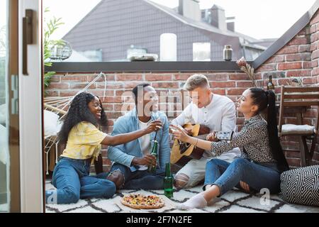 Groupe de jeunes gens divers assis sur le toit avec des cocktails et de la pizza. Des amis joyeux qui utilisent la guitare pour se détendre pendant leur temps libre. Banque D'Images