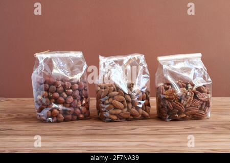 Trois types de noix - noisettes, amandes, noix de pécan - en sachets transparents sur une table en bois contre un mur brun. Gros plan Banque D'Images