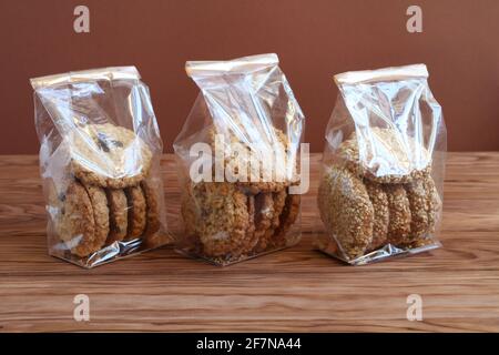 Trois types de biscuits d'avoine - avec chocolat, noix et raisins secs, et graines de sésame - dans des sacs transparents sur une table en bois contre un mur brun. C Banque D'Images