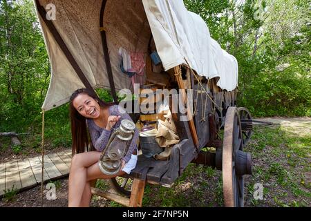 Femme asiatique excitée avec lampe à huile rétro regardant l'appareil photo et souriant tout en étant assis sur un vieux wagon pendant la visite à fort Edmonton en Alberta, Canada Banque D'Images