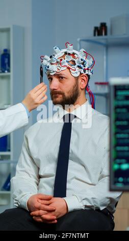 Homme assis sur une chaise neurologique avec un micro-casque à balayage d'ondes brainables tandis que le chercheur examine l'état de santé écrit sur le presse-papiers. Médecins travaillant dans un laboratoire d'étude du cerveau avec des moniteurs montrant la lecture EEG. Banque D'Images