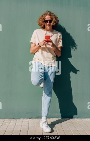 portrait vertical complet d'un jeune homme du caucase utilisant un smartphone rouge. Il se repose sur un mur vert et sourit. Jour ensoleillé projetant des ombres profondes Banque D'Images