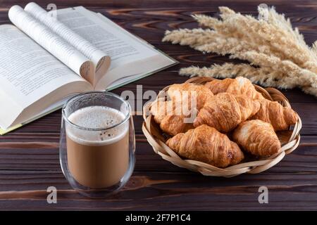 Croissants et un verre de café au lait sur une table en bois brun foncé. Boisson chaude et livre ouvert sur les planches. Concept petit déjeuner français, pause café Banque D'Images