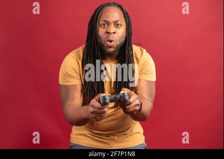Homme d'inspiration afro-américaine passionné de noir avec de longues dreadlocks, vêtu de vêtements décontractés, jouant dans un jeu vidéo en ligne, debout sur fond rouge, en utilisant le jeu de joystick consol, en s'amusant seul Banque D'Images