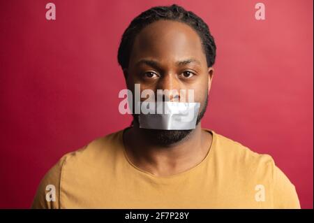 Le concept de racisme avec un homme africain en colère avec une bouche scellée avec la bande regardant la caméra, isolé sur fond rouge, sans voix, a énervé Guy en raison de l'incapacité de dépasser son opinion Banque D'Images