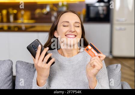 Photo d'une jeune femme excitée qui fait des achats en ligne tant attendus, heureuse femme tient un smartphone et une carte de crédit, magasiner en ligne, rêver avec les yeux fermés au sujet de nouvelles choses. Concept de consumérisme Banque D'Images