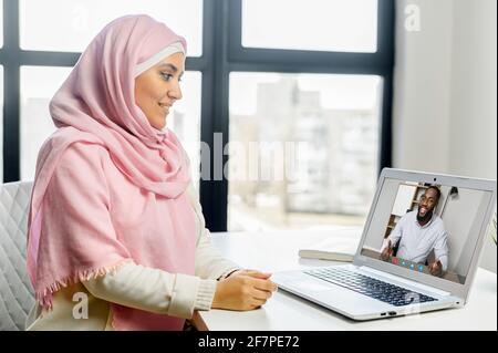 Jeune femme arabe musulmane conseiller d'affaires dans hijab regardant l'écran parler au jeune homme noir, ayant la consultation virtuelle avec le client, session vidéo en ligne ou de réunion via ordinateur portable du bureau Banque D'Images
