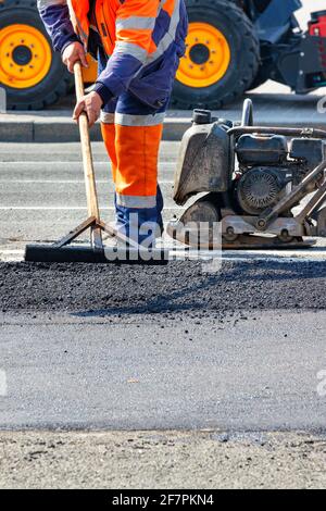 Un travailleur routier répare une section de route avec un niveau de bois devant un rouleau, un rameur et une niveleuse. Image verticale, espace de copie. Banque D'Images