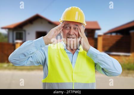 Homme de constructeur âgé avec choqué surpris l'expression submergée faisant le geste touchant les temples sur le fond du bâtiment de la maison Banque D'Images