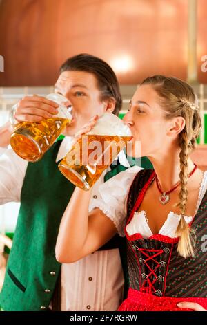 Jeune homme et femme dans le traditionnel Tracht avec verre à bière dans la brasserie, en face de la bouilloire de brassage Banque D'Images