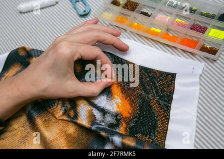 Processus de broderie de perles. Gros plan - les mains des femmes enfilent des perles sur une aiguille et les coudre sur le tissu. La femme aime la broderie. Banque D'Images