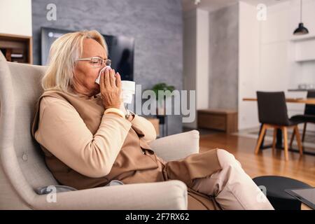 Une femme sénior malade assise sur une chaise à la maison et soufflant son nez. Il a attrapé le froid. Banque D'Images