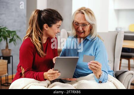 Mère et fille surjoyeuse et excitée assise à la maison ensemble et utilisant une tablette pour faire du shopping en ligne. Fille tenant un comprimé tandis que mère tenant un cré Banque D'Images