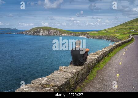 Homme d'âge moyen assis sur un mur, en admirant la vue incroyable de Slea Head, Coumeenoole Beach et Dingle Peninsula partie de Wild Atlantic Way, Kerry, Irlande Banque D'Images