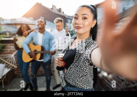Adorable jeune femme prenant le selfie tandis que ses amis drôles jouant de la guitare et chantant en arrière-plan. Quatre personnes de course mixte traînaient ensemble sur le toit. Banque D'Images