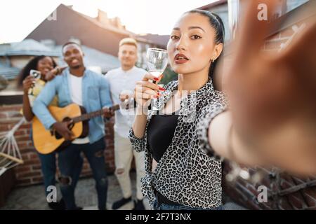 Adorable jeune femme prenant le selfie tandis que ses amis drôles jouant de la guitare et chantant en arrière-plan. Quatre personnes de course mixte traînaient ensemble sur le toit. Banque D'Images