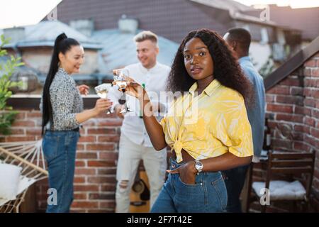 Femme afro-américaine souriante debout sur le toit avec une coupe de champagne. Trois amis multiculturels discutant et buvant derrière eux. Les jeunes se détendent lors d'une fête. Banque D'Images