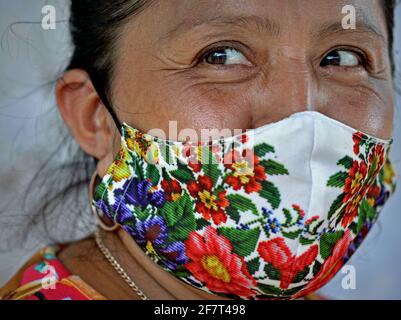 Une femme mexicaine âgée porte un masque en tissu folklorique imprimé coloré pendant la pandémie mondiale du coronavirus et sourit avec ses yeux timides. Banque D'Images