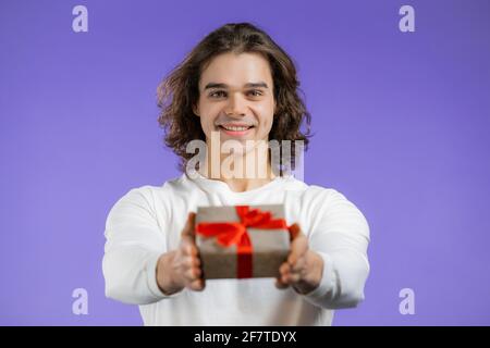 Charmant homme donne boîte cadeau à la main à l'appareil photo sur fond de mur violet. Guy souriant, il est content du présent. Portrait de studio Banque D'Images