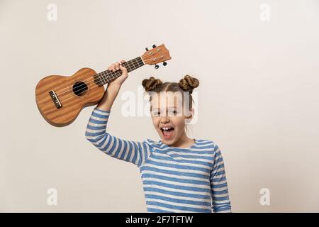 La fille chante et joue le ukulele. L'enfant rit, pose pour l'appareil photo et aime la musique. Apprendre à jouer ukuleles Banque D'Images