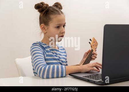 Une adolescente apprend à jouer à l'ukulele et regarde dans son ordinateur portable. Banque D'Images