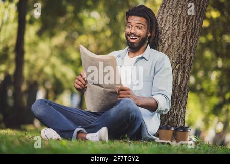 Photo pleine grandeur de brunette excitée afro américain homme assis grass park lit le journal stupéfié dehors dans le week-end extérieur Banque D'Images