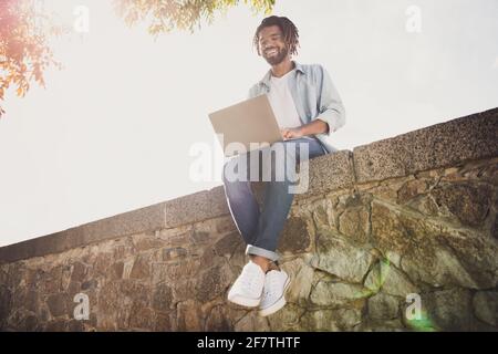 Portrait de sympathique gai focalisé funky gars spécialiste informatique voyageur assis sur un mur en pierre à l'aide d'un ordinateur portable, jour ensoleillé à l'extérieur Banque D'Images