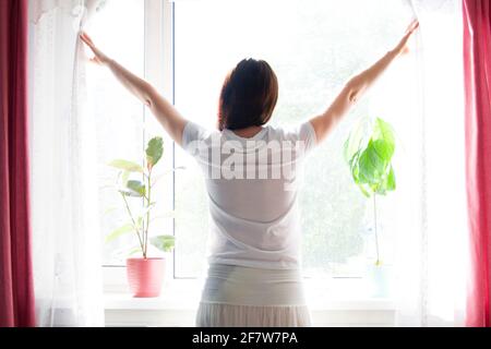 Une femme ouvre les rideaux le matin pour laisser entrer beaucoup de soleil. Banque D'Images